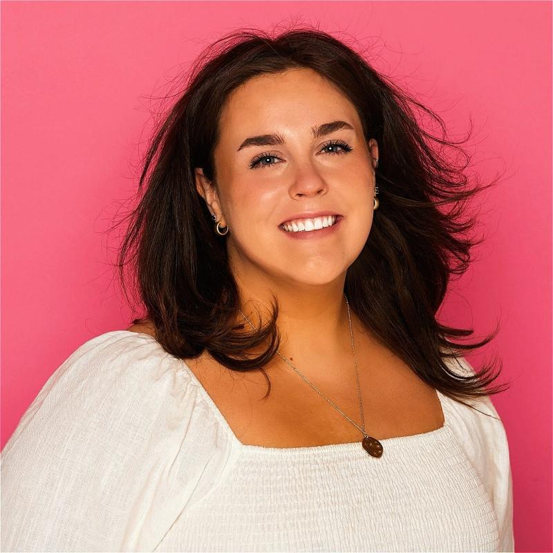 Portrait of a smiling woman with shoulder-length brown hair, wearing a white blouse and a pendant necklace, against a pink background.