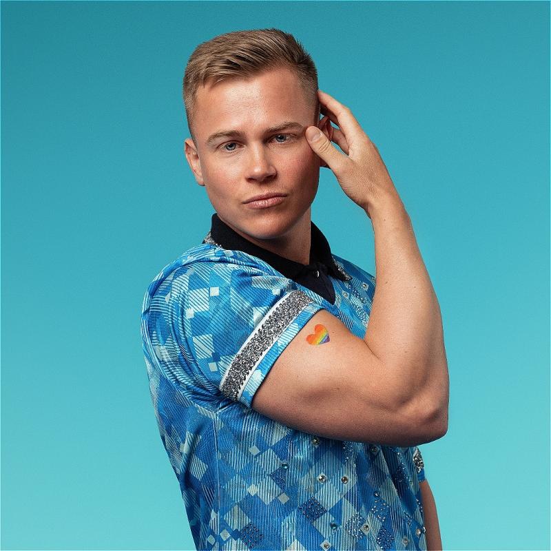 Young man with short blond hair, wearing a blue patterned shirt, poses against a teal background. His right hand is touching his temple, and there is a rainbow heart sticker on his left arm.