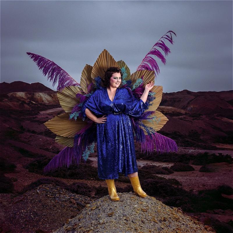 A woman in a sparkling blue dress, adorned with large golden and purple fans and feathers, stands on a rocky hill with a dramatic overcast sky and a rugged landscape in the background. She wears yellow boots and appears joyful as she poses with one hand raised.