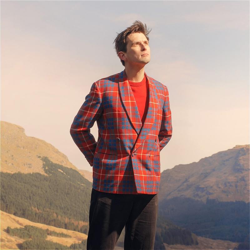 Man in a red and blue plaid blazer and black pants standing on a hill with mountainous terrain in the background, looking upwards with a serene expression.