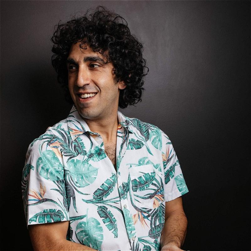 Portrait of a smiling man with curly hair looking to the side, wearing a white tropical print shirt against a dark background.