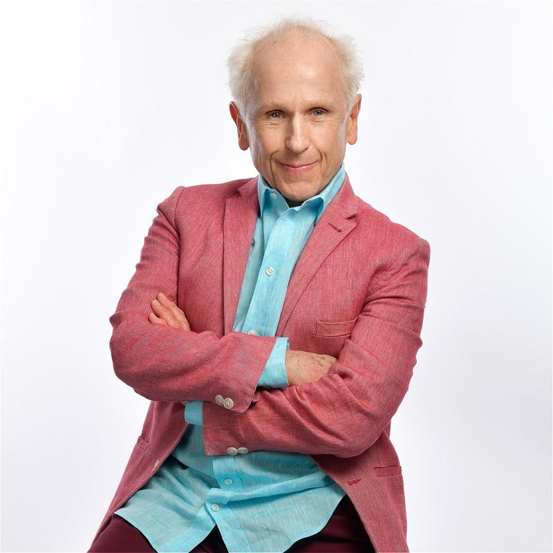 Elderly man with white hair posing confidently on a stool, wearing a coral blazer, turquoise shirt, and maroon trousers.