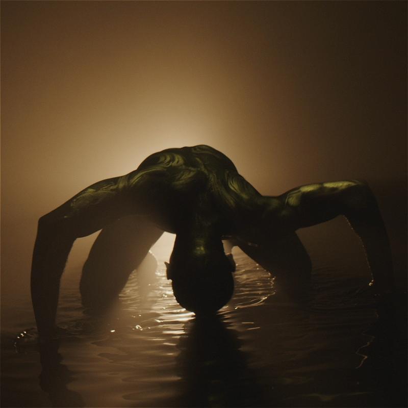 A muscular human figure is bending forward in shallow water, illuminated dramatically from behind by a golden light that casts a silhouette and reflects on the water's surface. The setting appears moody and artistic, emphasizing the contours and textures of the figure's back and arms.