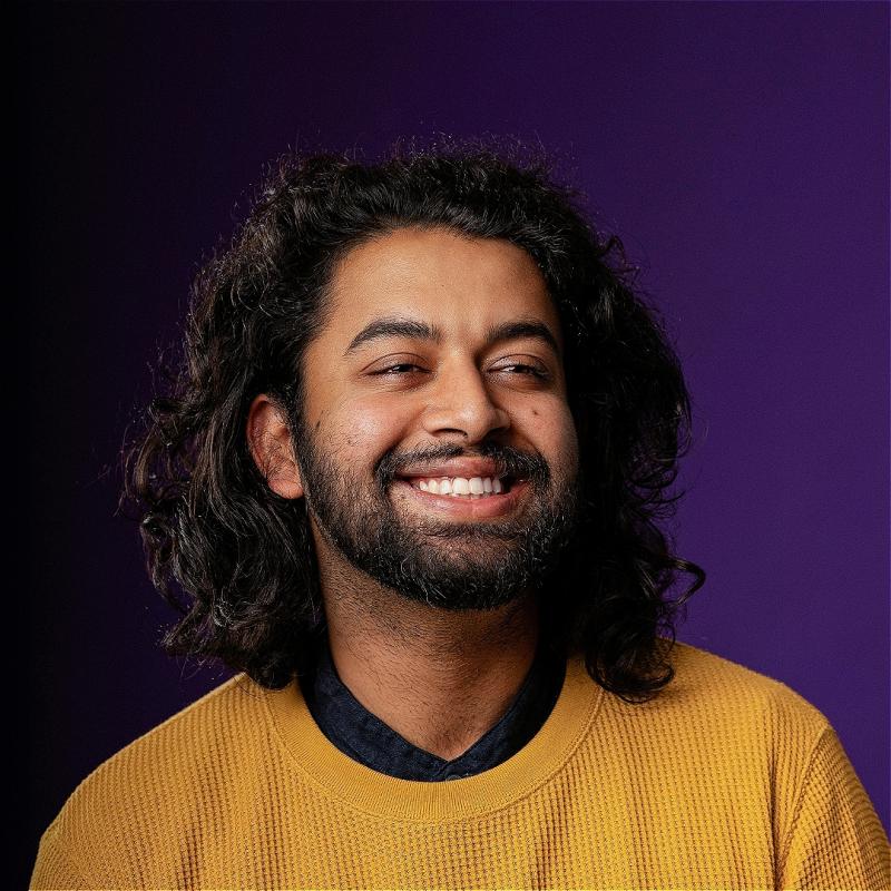 Portrait of a joyful man with long curly hair, wearing a yellow sweater over a blue shirt, smiling broadly against a purple background.