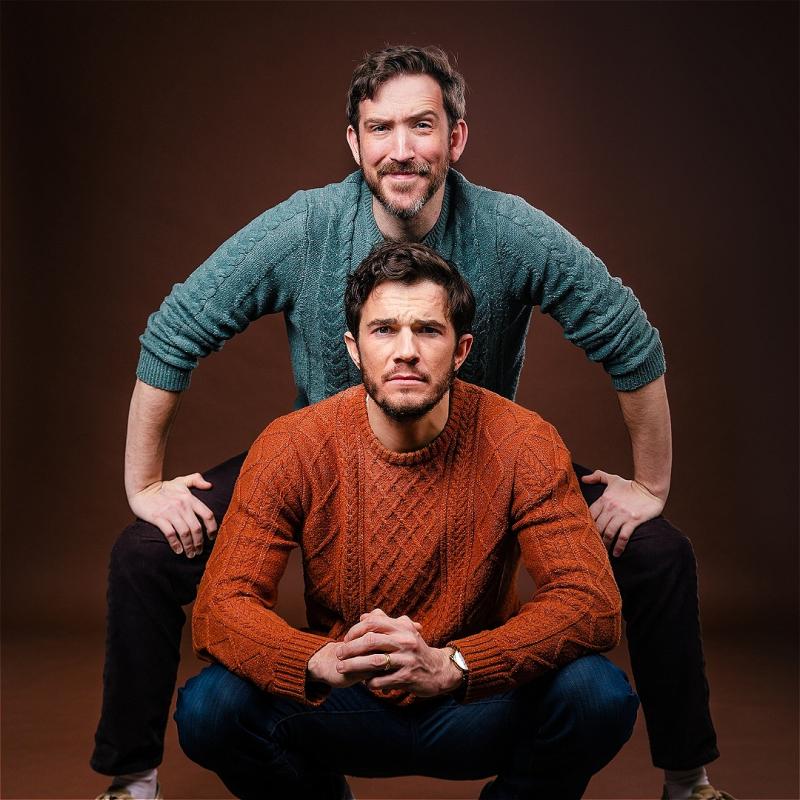 Two men posing in a studio, with one crouching far down and the other straddling behind him, both wearing knitted sweaters and jeans, set against a brown backdrop. The crouched man wears an orange sweater and white sneakers, while the straddling man wears a green sweater and beige sneakers, both smiling slightly at the camera.