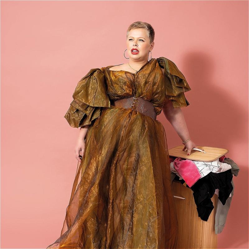 Laura stands in front of a pink background in a dramatic brown ball gown. She has on large hooped earrings and it staring into the distance. She is leaning on an overflowing laundry bin. 