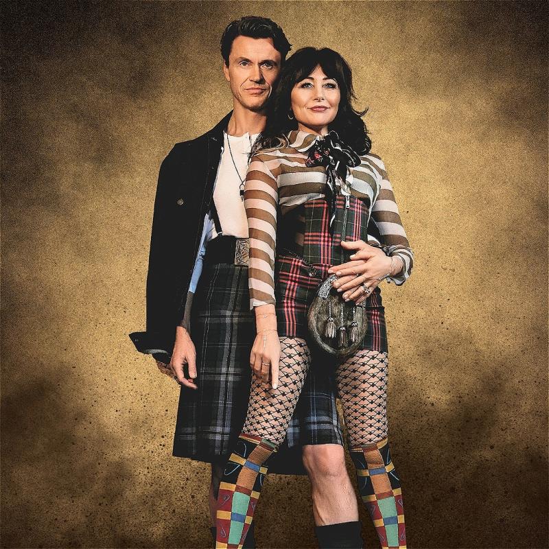 A man and a woman dressed in avant-garde Scottish-inspired outfits including tartan kilts, patterned fishnet stockings, and colorful mismatched socks, standing closely together against a textured golden background.