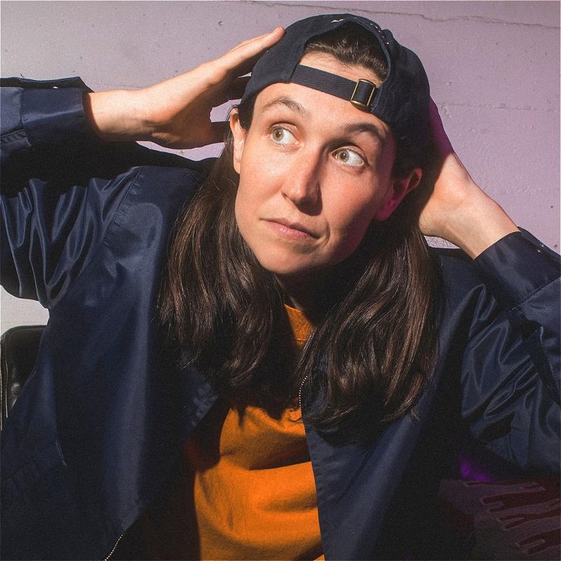 A young woman with long brown hair wearing a backwards baseball cap, navy blue bomber jacket, and orange t-shirt, sitting with a playful expression and hand on her head against a light purple background.