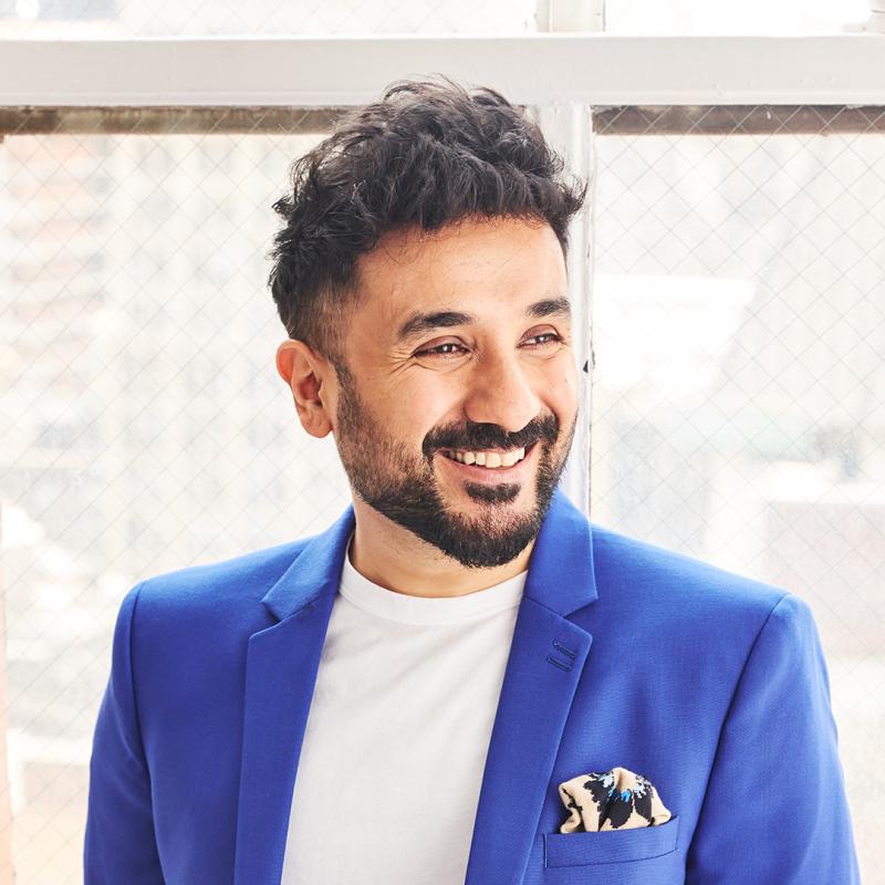 Vir Das looks smilingly off camera and wears a blue suit with a white shirt.