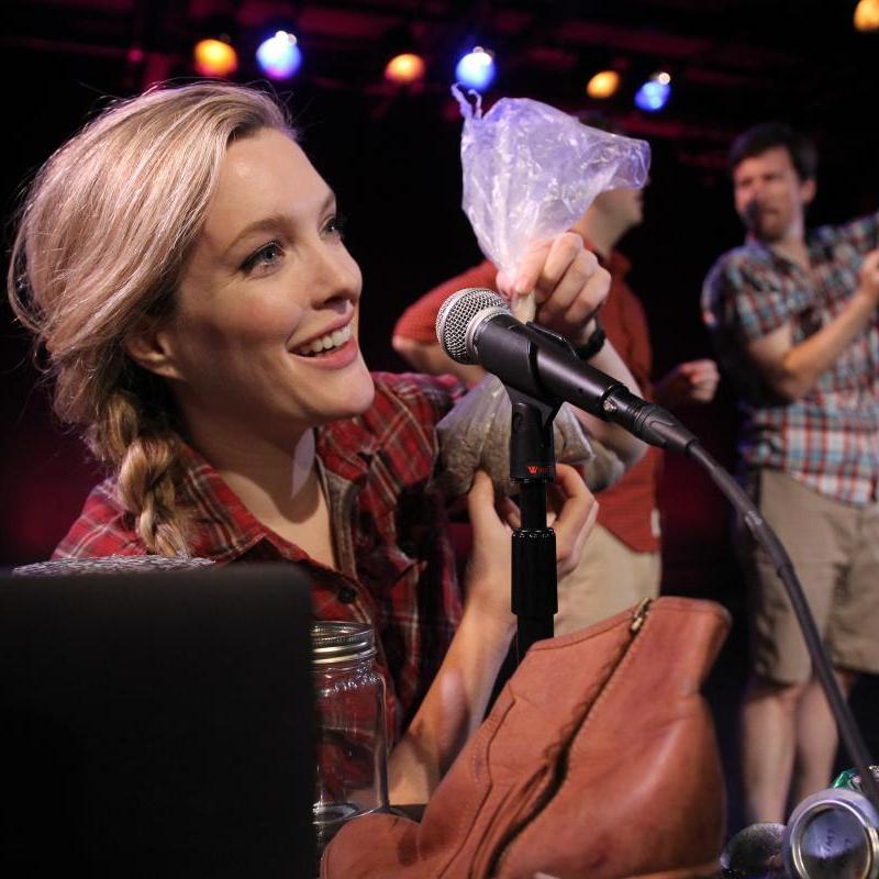A Foley artist holds a plastic bag and makes noises into a microphone. Two men are blurred in the background.