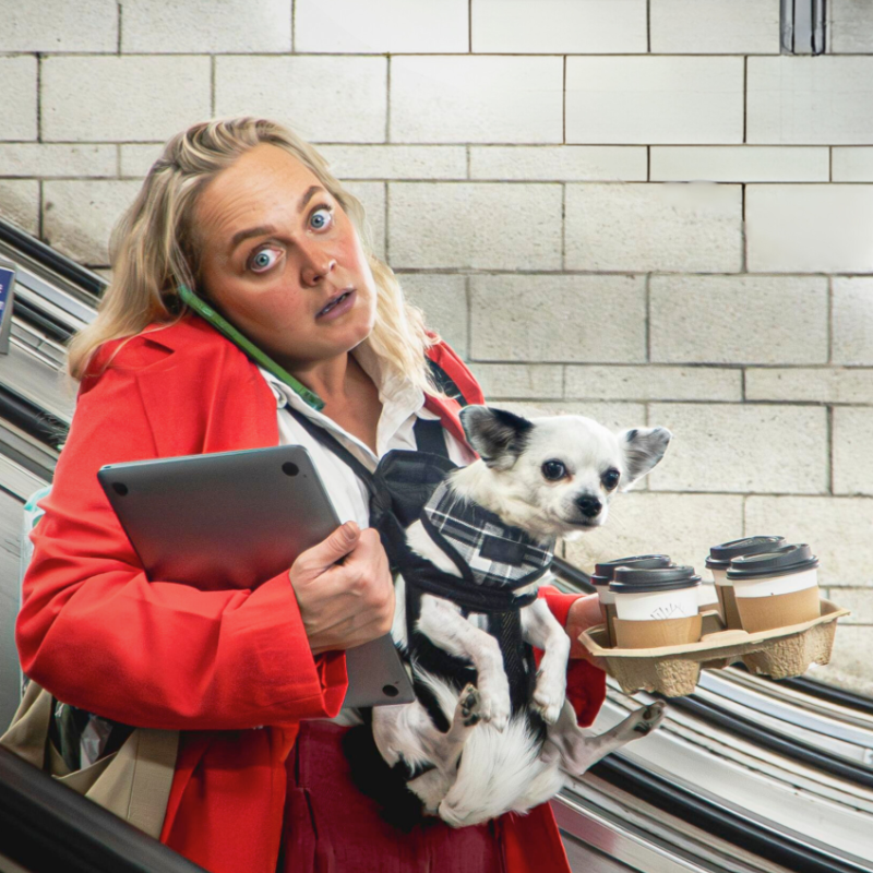 A busy looking woman in a red suit and corporate wear stands on an escalator holding a laptop, dog in a harness and four takeaway coffee cups in a tray.