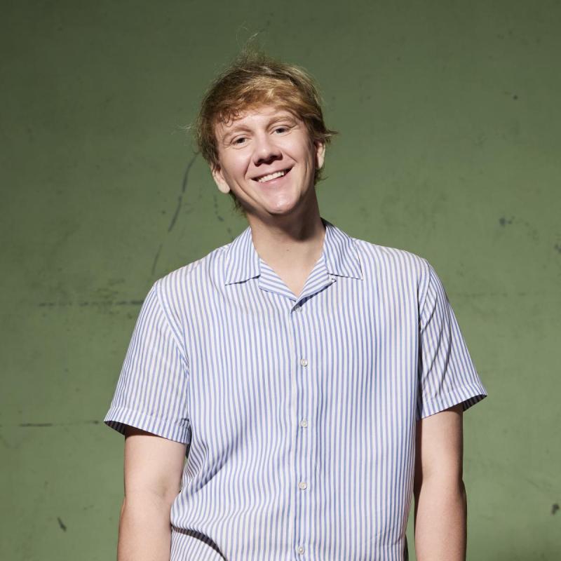 Smiling photo of the performer in a striped shirt standing against an olive green backdrop.