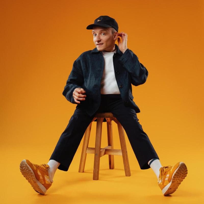 Will Robbins sat on a stool against an orange background with orange trainers and cap.