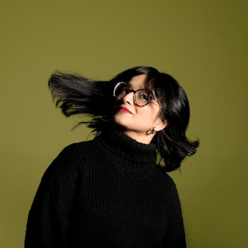 Headshot of Shalaka flipping their hair. Performer wears a black polo neck jumper and red lipstick.