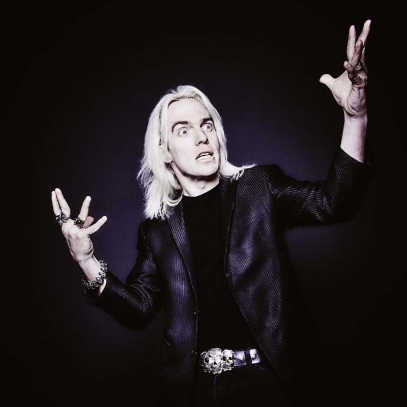 John Robertson wearing all black against a black background gesticulating wildly with a crazed look on their face