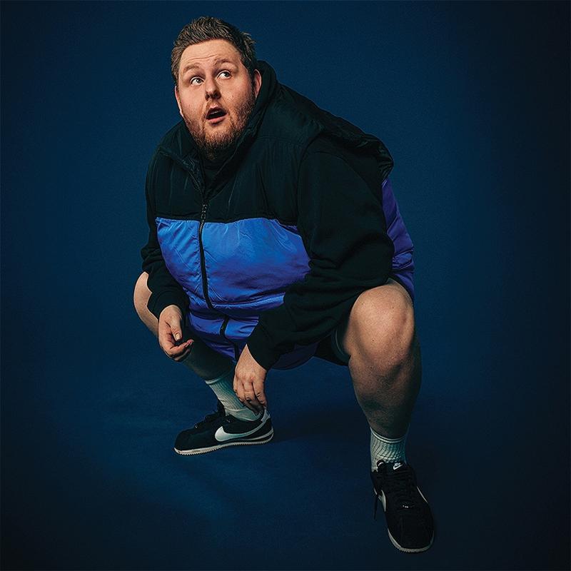 A large man squats down against a blue background wearing a blue puffer gilet, he looks agape up to the right corner of the image.