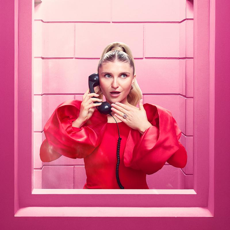 Sophie in a bright pink dress holds a phone standing behind glass.