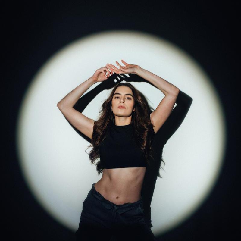 Amelia poses with her arms above her head in a black crop top - there is a front spotlight on her