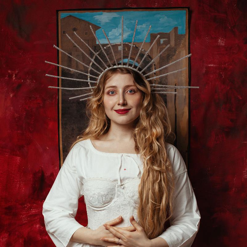 Bebe Cave wearing a long white dress and a headpiece gazes submissively into the camera.
