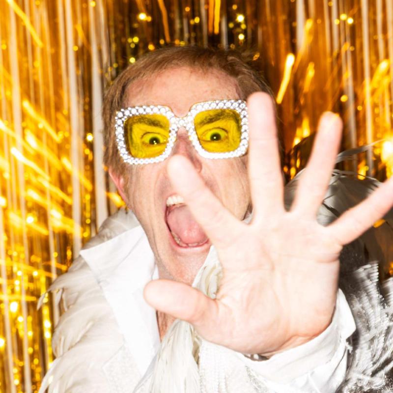 Image of man in white feathers and orange lensed sunglasses holding his hand up to the camera whilst shouting menacingly