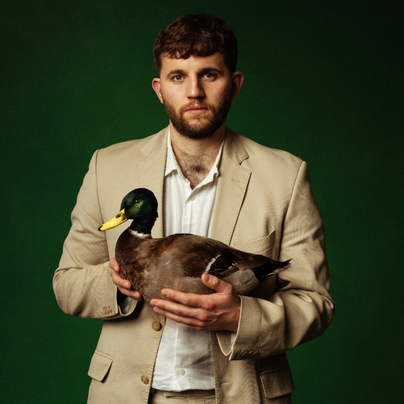 Rory stands in a beige suit, holding a live duck.