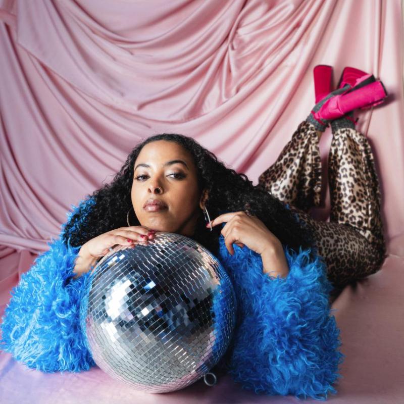 Performer lounges on top of a disco ball in a blue feather cardigan, leopard print leggings and heels.