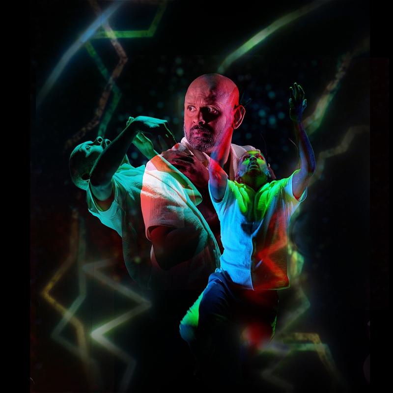 Ethereal image of a stocky bald man looking pensively over his shoulder whilst two figures dance expressively. The three men all blend together with green and red lights.