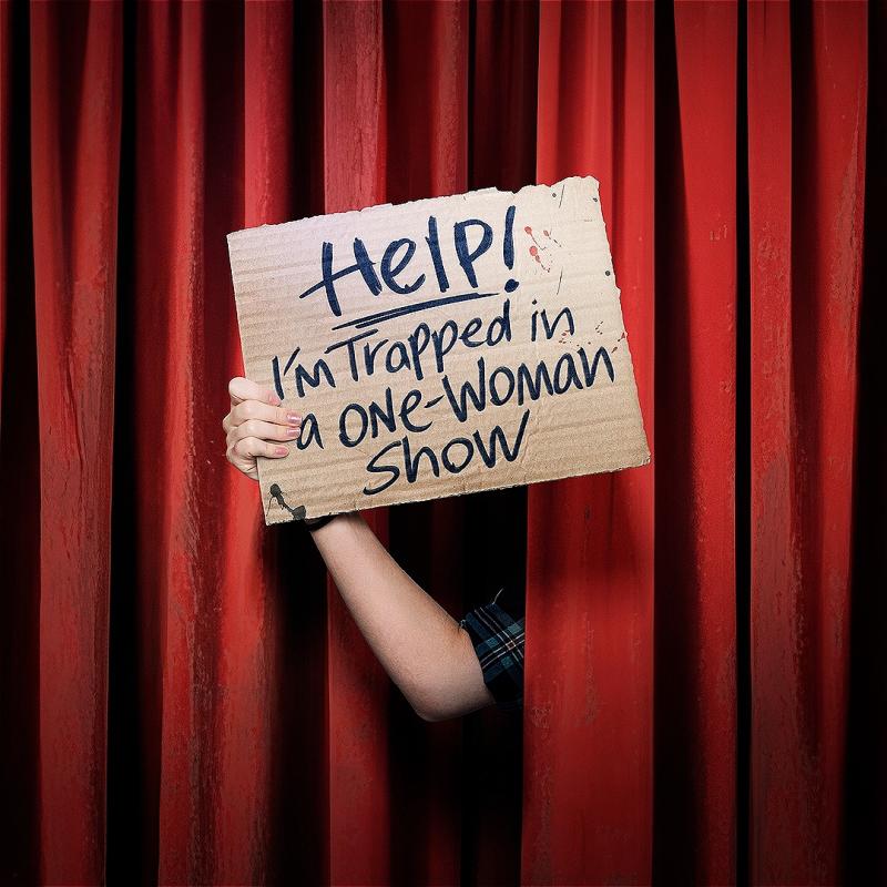 A hand appears holding a sign reading 'HELP! I'M TRAPPED IN A ONE WOMAN SHOW' from in between a red theatre curtain.
