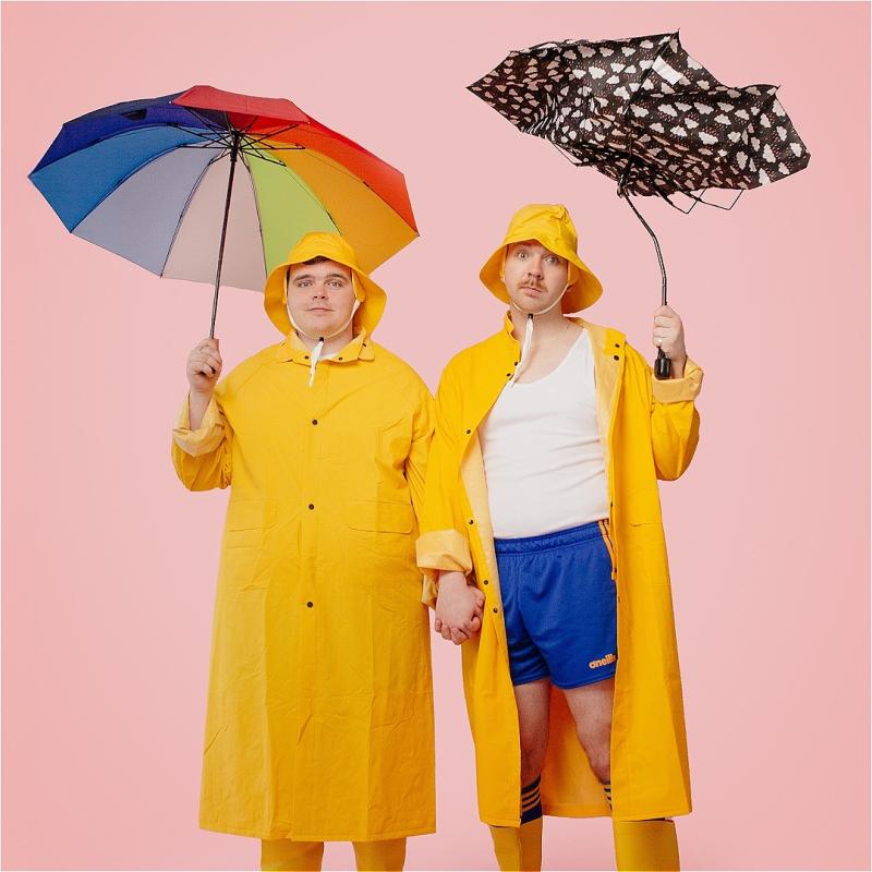 Two performers with hands joined are holding up umbrellas wearing yellow coats and matching fisherman hats.