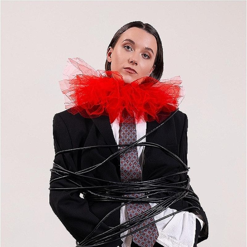 Claire Noy stands in a suit, tie and large red ruff looking particularly apathetic about the fact somebody has wrapped their microphone cable around them twenty times. They hold the microphone laxly in their right hand.