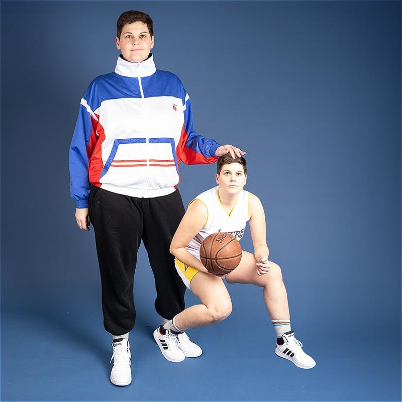 Performer Abby stands in a red white and blue tracksuit top and black trousers with their hand on top of a smaller version of themselves playing basketball.