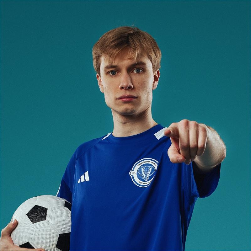 Ted points at the camera, holding a football in his other hand and wearing the royal blue jersey of Crudchester United