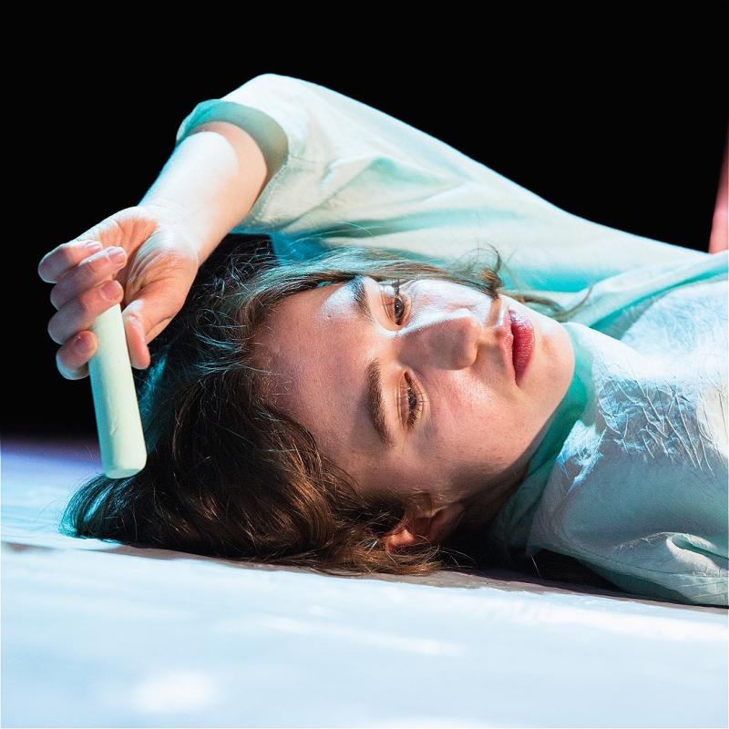Woman lays on the floor looking disheveled and holding a white cylinder