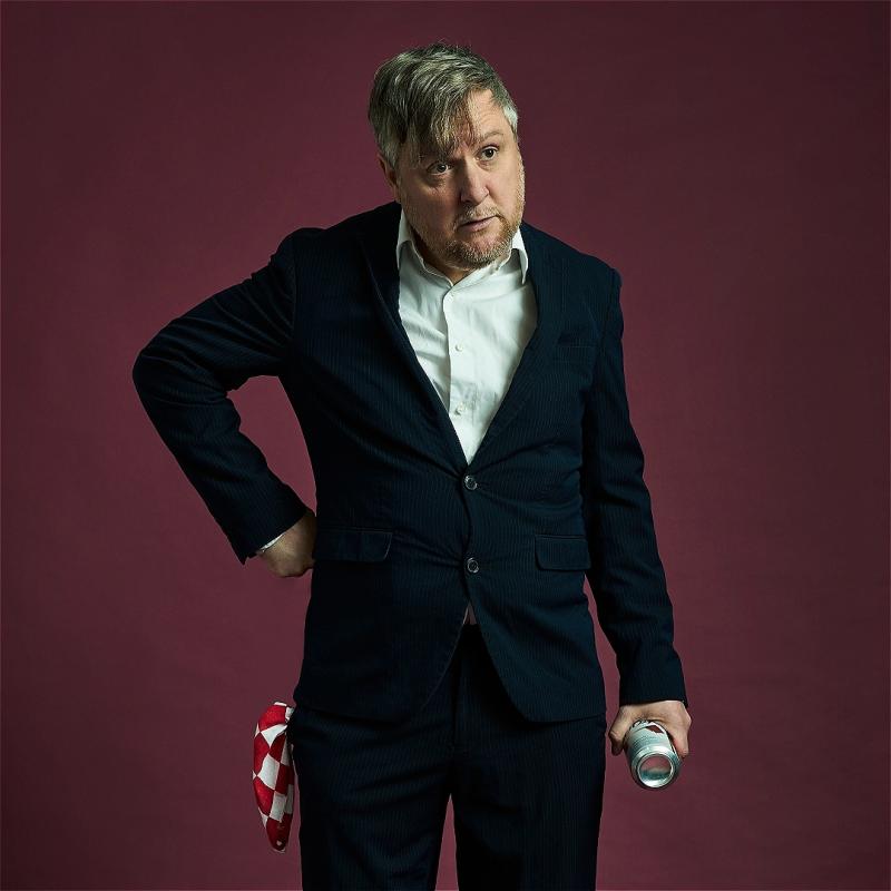 Tim Key stands in a suit with his hand on his hip against a purple backdrop