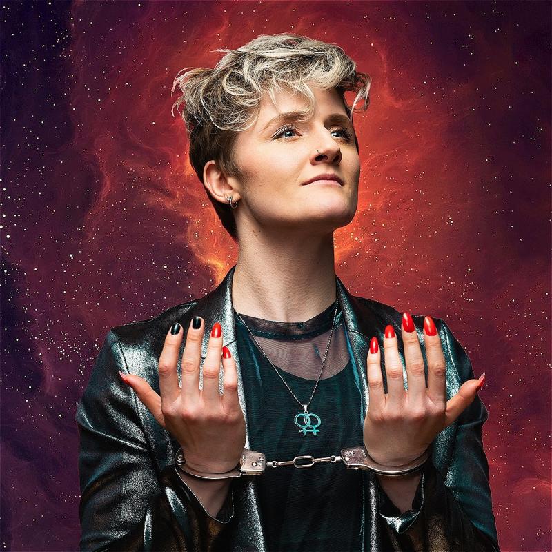 Performer stands against an intergalactic starry background with their hands in handcuffs.