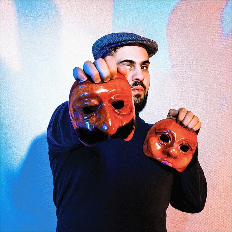 A performer wearing a cap holds up two theatre masks with one arm outstretched.
