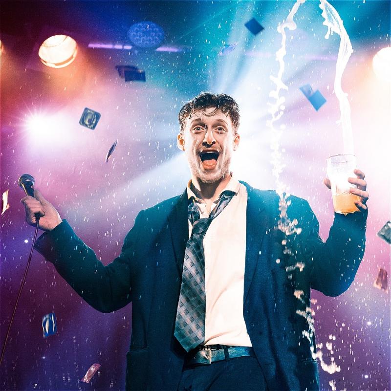 Jack Tucker stands brightly, slack-jawed and askew, holding a microphone and lager beer - which spills violently into the air.