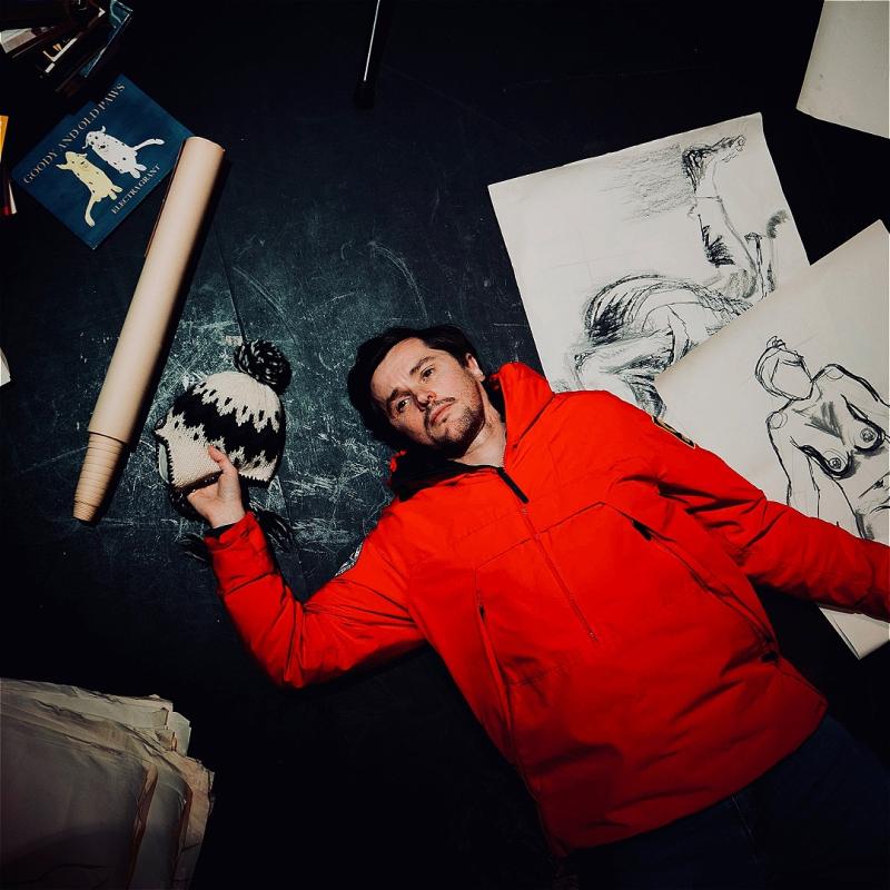 A man in a bright red jumper lays down next to line drawings of an exposed women, clutching a woolen trapper hat in his right hand