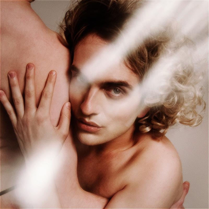 Blonde shirtless man holds on to another person as he stares at the camera, light flare over them.