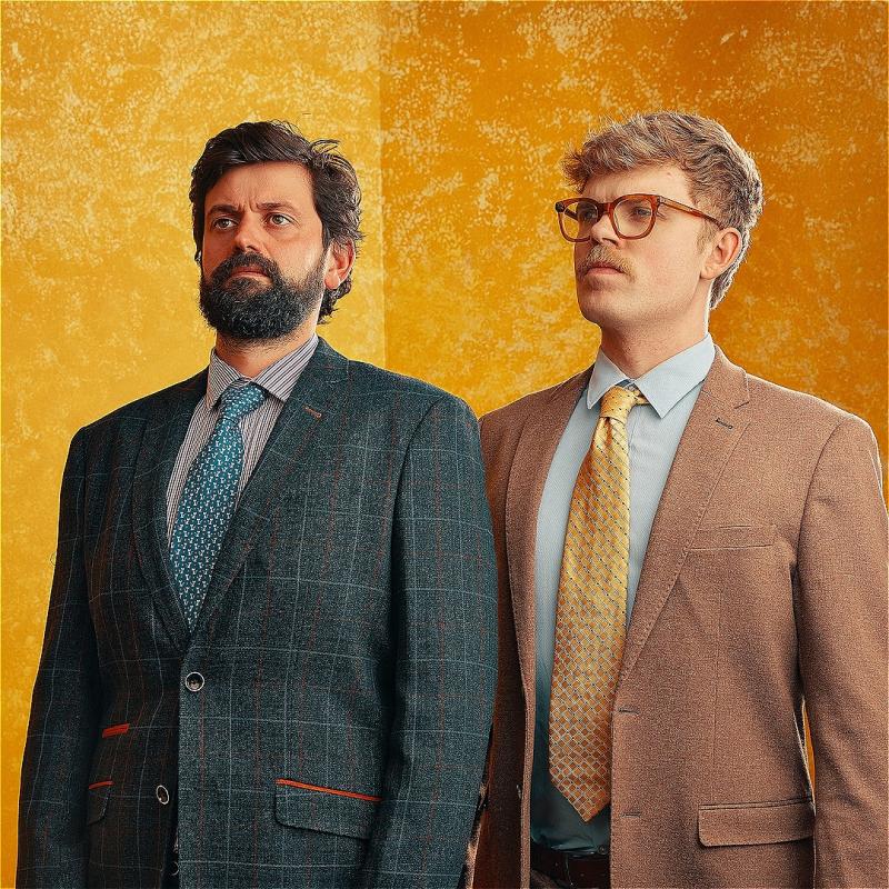 Fin Taylor and Horatio Gould stand next to one another wearing contrasting suit colours and looking serious.