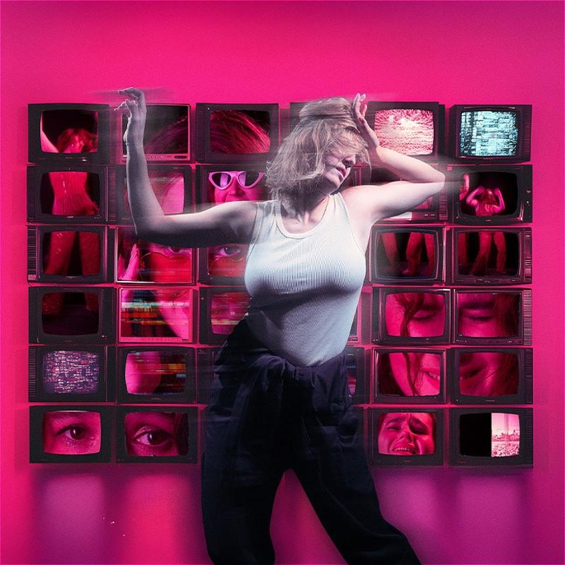 A woman dances chaotically in front of a 6x6 wall of TV screens, depicting another woman's face in a variety of positions against a hot pink backdrop.