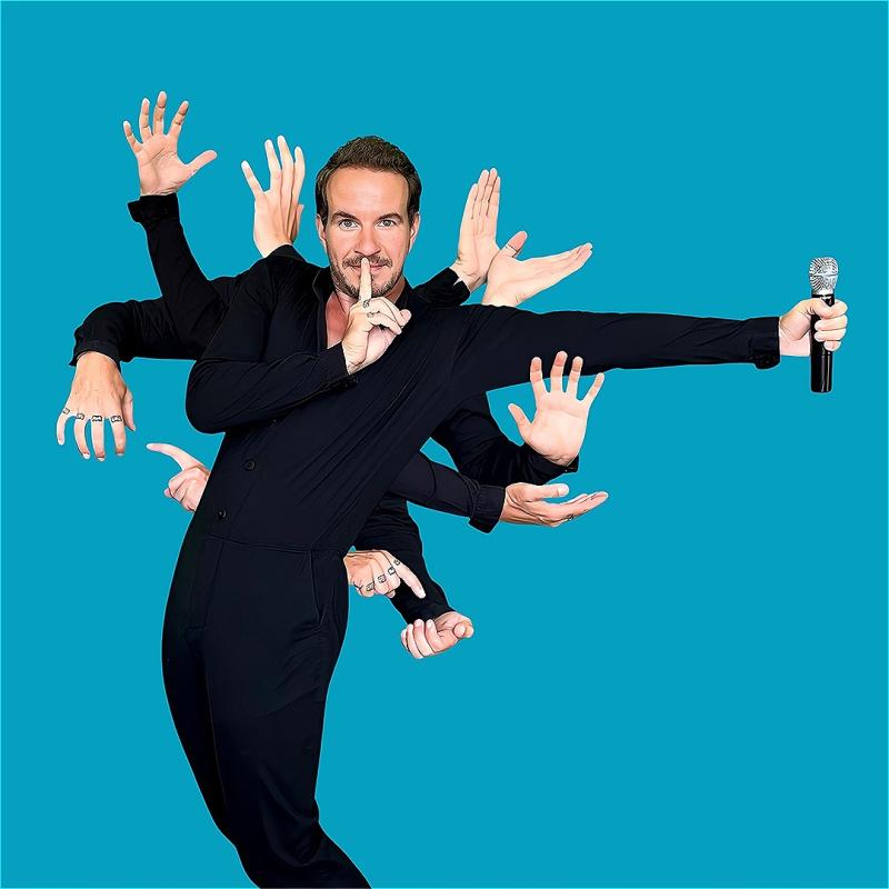A man dressed in black holds his finger to his lips in a shushing manner whilst eleven hands sprout from his body in an assortment of different positions. The hand stretched furthest from the man holds a microphone.