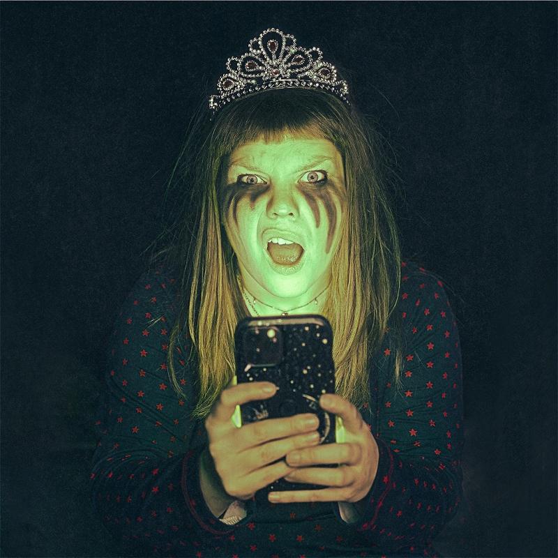 A ghastly ghoul lady stands with tiara on head looking perplexed by what she sees on her mobile phone which is planted in front of her face.