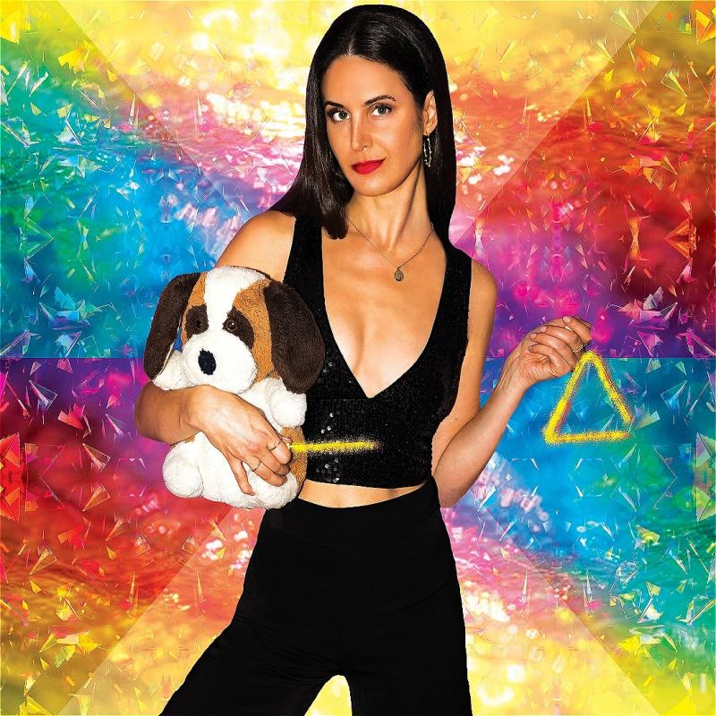 A woman stands against a rainbow backdrop holding a stuffed toy dog in one arm and a glittery triangle in her other hand.