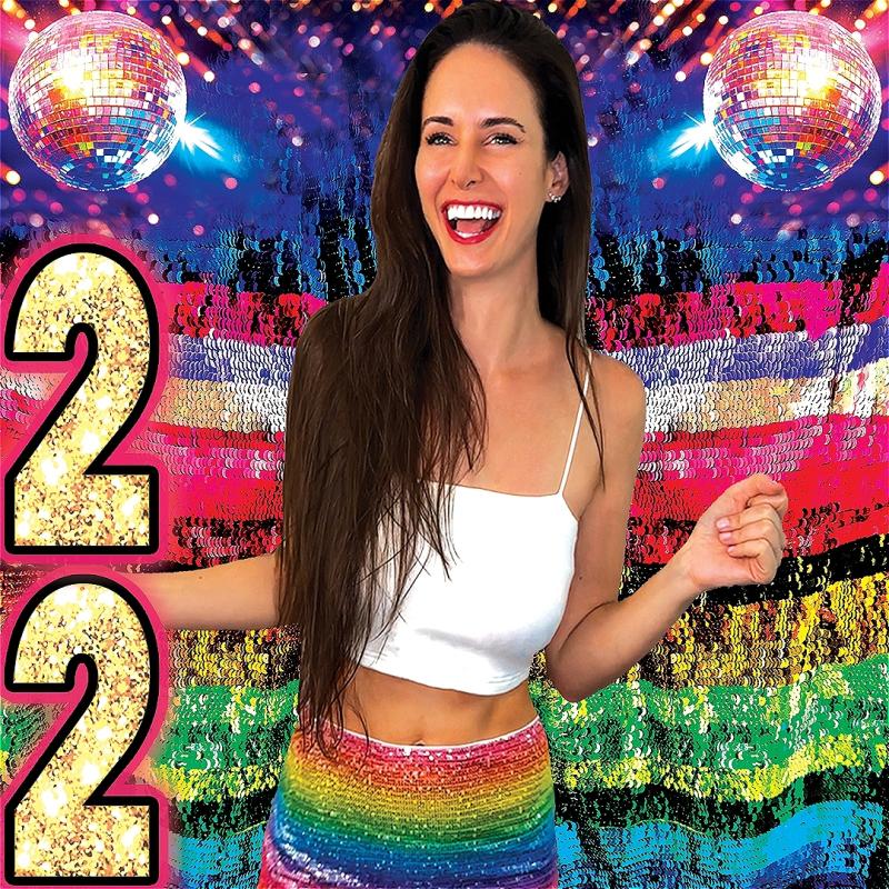 Brooke stands in front of an extremely glittered backdrop filled with colour, disco balls, patriotic red and blue, and the numbers '2' and '2' which are stacked on top of each other in the bottom left corner.
