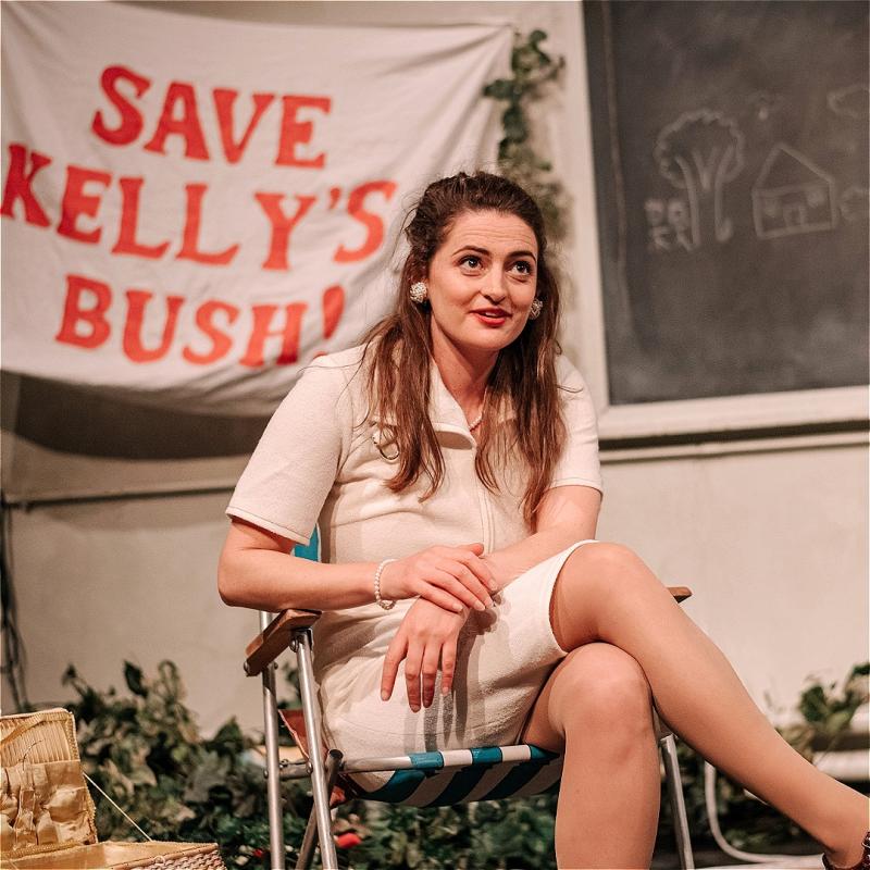 A performer with long brown hair sits cross-legged on a deckchair. A banner behind them says 'Save Kelly's Bush!'