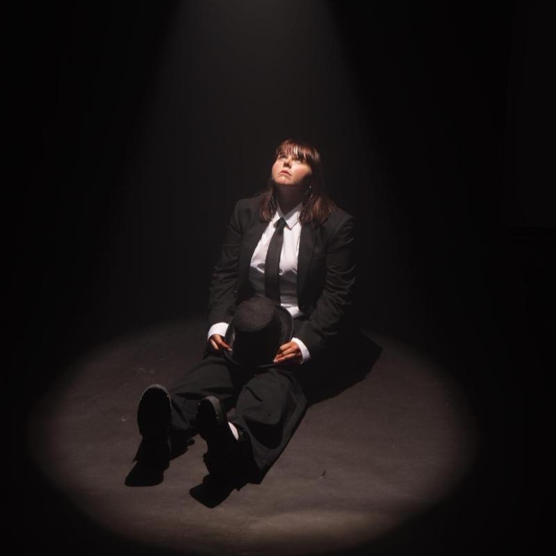 A performer dressed in a black suit and tie sits on the floor, a spotlight shining on them. Their legs are stretched out in front of them, they're holding a hat on their lap, and they are staring up at the spotlight.