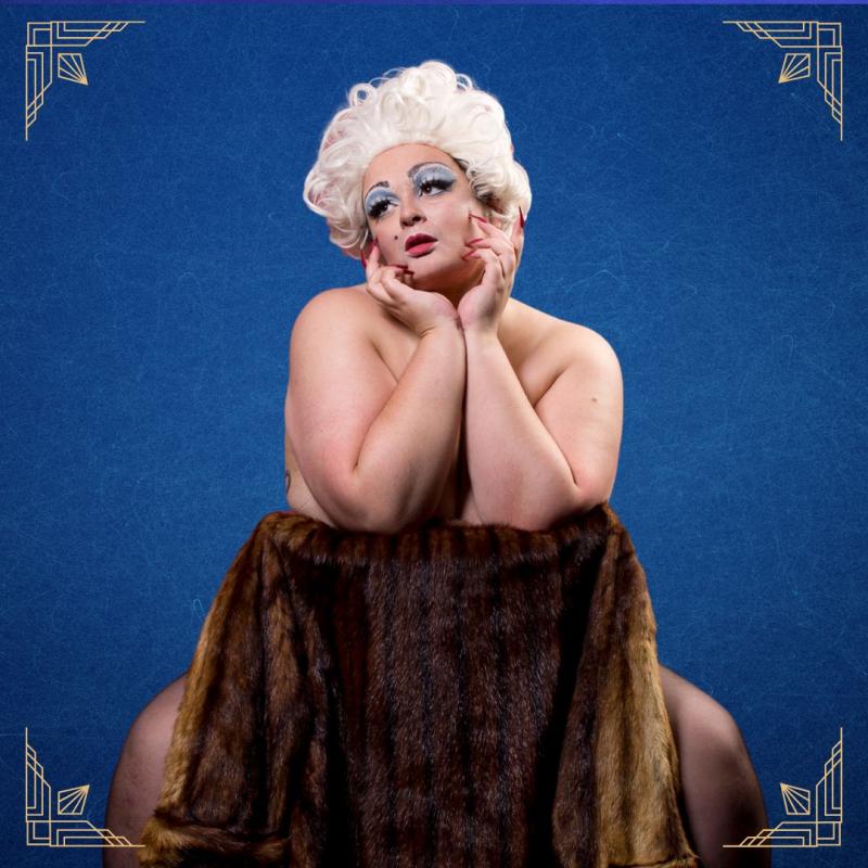 A performer in a marilyn monroe style blonde wig straddles the back of a chair, facing the camera. they appear to be tastefully nude. the background is a deep blue, and each corner of the image is decorated with an art deco-esque flourish