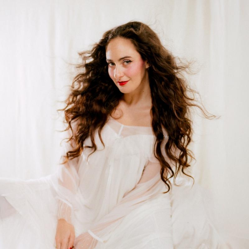 Cat Cohen sitting and smiling sweetly in an angelic white gown against a white fabric backdrop