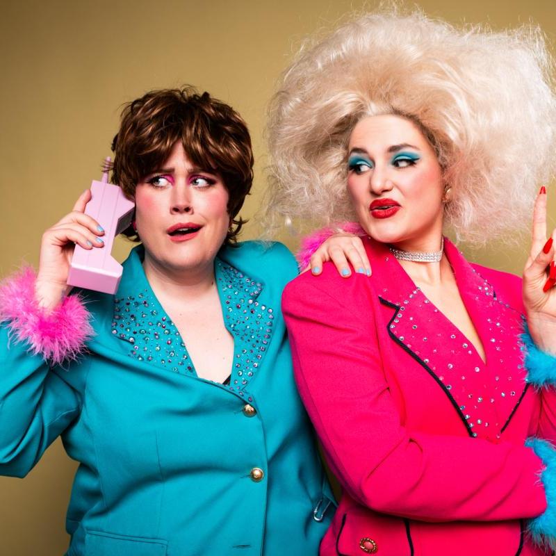The performers in garish big blue and pink 80s style suits and wigs, one holding a vintage pink phone